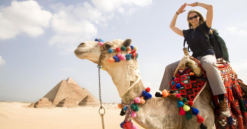 Giza Cairo pyamids background female tourist making shape of pyramids with arms while sitting on camel e1715087286193 1